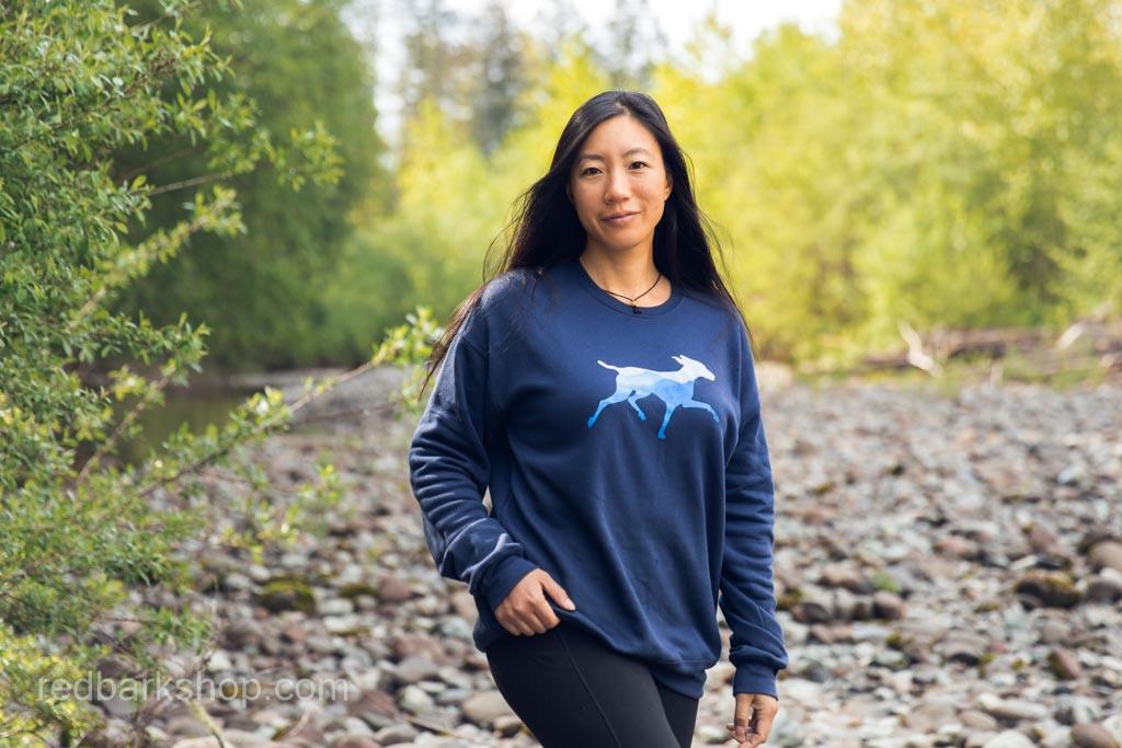 an Asian girl stands in front of a rocky beach wearing a navy mountain dog sweater in Squamish